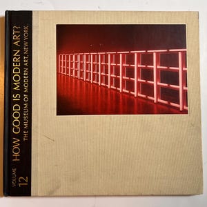 May include: A hardcover book titled "How Good Is Modern Art?" Volume 12, from The Museum of Modern Art, New York. The cover features a photograph of a red-lit installation. The book has a beige cover and a black spine.