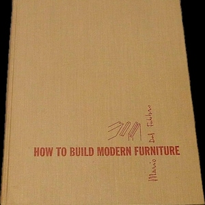 May include: A tan-colored book titled "HOW TO BUILD MODERN FURNITURE" in red lettering. The author's names, Mario and Daf Fabro, are written vertically in red ink. The book cover has a simple, minimalist design.