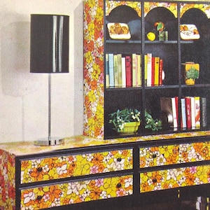 Puede incluir: Una estantería negra con seis estantes, decorada con un papel tapiz floral en tonos amarillos, naranjas y blancos. La estantería tiene una sección superior con tres estantes y una sección inferior con tres cajones. Los cajones también están decorados con el mismo papel tapiz floral. Una lámpara negra con una pantalla negra está en el lado izquierdo de la imagen.