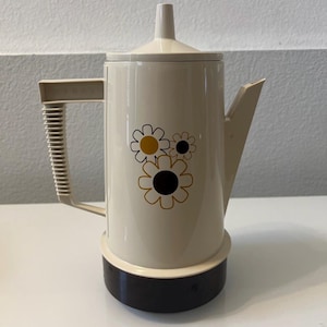 May include: A cream-colored electric percolator coffee pot with a dark brown base and a matching lid. The body features a graphic print of three stylized flowers in gold and dark brown. The handle is ribbed and embossed with the word REGAL.