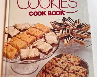 Könnte beinhalten: Ein Kochbuch mit dem Titel "Better Homes and Gardens Homemade Cookies Cook Book" mit verschiedenen Keksen. Die Kekse sind auf Glas- und Silberplatten angeordnet. Die Kekse sind in Braun-, Beige- und Weißtönen gehalten.