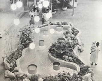 May include: Black and white photograph of an indoor shopping mall with a water feature. The water feature is surrounded by plants and rocks. People are walking around the mall. Overhead, there are hanging globe lights.