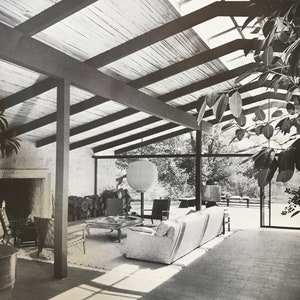 以下が含まれることがあります： 白黒写真のミッドセンチュリーモダンなリビングルーム。傾斜した木製の天井、天窓、暖炉、自然の風景を望む大きな窓があります。 家具には、ソファ、椅子、コーヒーテーブルが含まれています。