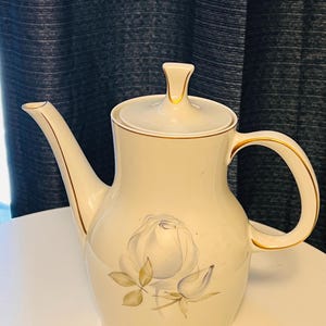May include: Cream-colored ceramic teapot with a floral rose design. The teapot has a curved spout, a rounded body, and a lid with a small handle. Gold trim accents the edges of the spout, handle, and lid. A painted white rose with blue and green accents decorates the front.