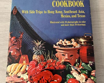 May include: A hardcover book titled Trader Vic's Pacific Island Cookbook with a blue cover featuring a colorful still life of tropical fruits, vegetables, a lobster, a ceramic bowl, and a statue against a dark background with text.