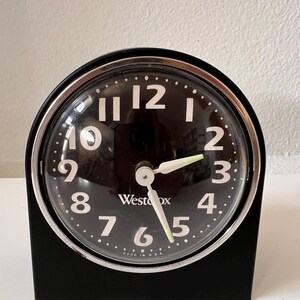 May include: A black Westclox analog alarm clock with a round face, white Arabic numerals, and a silver-toned bezel. The clock features white hour and minute hands with luminous tips, a thin white second hand, and the text Made in USA at the bottom.