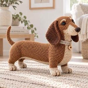 Puede incluir: Un perro salchicha de ganchillo hecho con hilo marrón y crema. El juguete tiene orejas largas y caídas, una cola corta y un collar gris claro con una pequeña etiqueta redonda de madera que presenta un corazón recortado. Ojos de plástico.