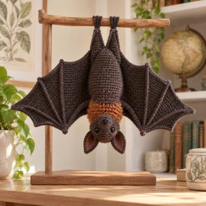 May include: A dark brown crocheted bat hanging upside down from a wooden stand. The bat features a textured body, wide outstretched wings, and a patch of lighter orange-brown yarn around its neck. It has dark eyes and pointed ears.
