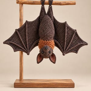 May include: A crocheted bat toy with dark brown wings and body, featuring a lighter orange-brown neck ruff and inner ears. The bat is hanging upside down from a wooden T-shaped display stand with a rectangular base, showcasing detailed yarn stitching.