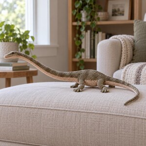 Könnte beinhalten: Ein gehäkeltes Dinosaurier-Spielzeug mit langem Hals und Schwanz, das einen strukturierten olivgrünen Rücken und eine hellbeige Unterseite aufweist. Die Kreatur hat vier Beine mit sichtbaren Zehen und einen kleinen Kopf mit dunklen Augen.