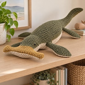 Peut inclure: Un jouet en peluche requin-marteau au crochet avec un dessus vert olive foncé, un dessous beige clair et une tête en forme de marteau jaune moutarde texturée. Le jouet possède de petits yeux en perles noires et une longue queue courbée.