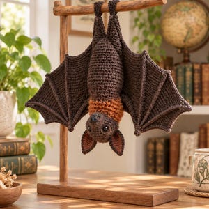 May include: A dark brown crocheted bat hanging upside down from a wooden T-shaped stand. The bat features a textured body, large spread wings with defined ribbing, a lighter orange-brown neck ruff, pointed ears, and small black eyes.