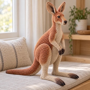 Puede incluir: Un juguete de canguro hecho a ganchillo con un patrón de puntos texturizado. El canguro tiene la espalda y la cola de color marrón rojizo, con el pecho y el vientre de color crema. Presenta detalles en marrón oscuro en las puntas de las patas.