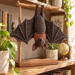 May include: A crocheted bat toy hanging upside down from a wooden T-shaped stand. The bat has dark brown wings and body, with a textured orange collar around its neck, black eyes, and pink-lined ears. The stand features a rectangular base.