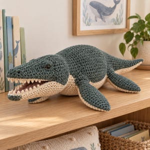 May include: A dark green and cream-coloured crochet plush toy shaped like a prehistoric marine reptile. The creature features a textured yarn surface, an open mouth with visible individual teeth, dark eyes, and four flipper-like limbs.