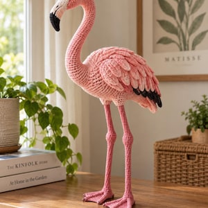 May include: A handcrafted crochet flamingo figurine featuring a soft pink body with layered wing details, long slender legs, and a curved neck. The beak is light pink with a black tip, and the eyes are small, dark, and round. The texture is yarn.