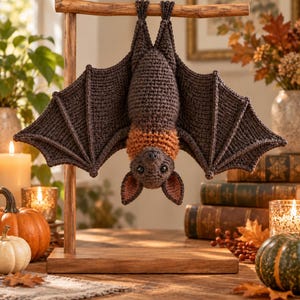 May include: A dark brown crocheted bat hanging upside down from a wooden T-shaped stand. The bat features large, textured wings, a lighter orange-brown neck ruff, pointed ears with pinkish inner details, and small black eyes facing forward.