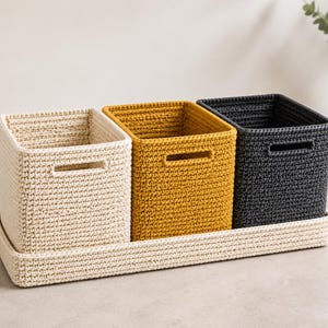 May include: Three square crochet storage baskets in cream, mustard yellow, and dark gray, each featuring a rectangular cutout handle. The baskets are arranged in a row and sit inside a long, rectangular cream-colored crochet tray.