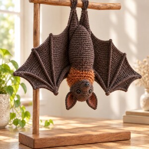 May include: A crocheted bat toy hanging upside down from a wooden T-shaped stand. The bat is primarily dark brown with a lighter orange-brown neck area, large pointed ears with pinkish interiors, and wide, textured wings with visible stitching.