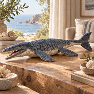 Peut inclure: Un jouet en peluche mosasaure crocheté présentant un dos texturé à motifs bleus et gris avec un ventre crème. La figurine de reptile marin a une bouche ouverte montrant de petites dents blanches, des yeux sombres et une longue queue.