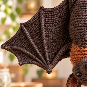 May include: A dark brown crocheted bat with a textured wing structure featuring prominent raised ridges. The bat has a fuzzy orange chest patch, a small dark nose, and ears with light pink inner detailing. The eyes are smooth, round, and black.
