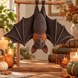 May include: A dark brown crocheted bat hanging upside down from a wooden T-shaped stand. The bat features large, textured wings, a lighter orange-brown neck ruff, pointed ears with pinkish inner details, and small black eyes facing forward.
