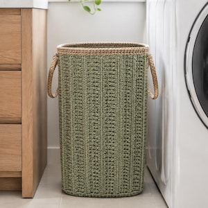 May include: A tall, cylindrical laundry basket featuring a textured, sage green crocheted body with vertical ribbed detailing. The basket has a reinforced tan rim and two matching tan braided handles attached to the sides for easy carrying.