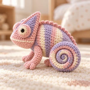 Op de afbeelding: Een gehaakt kameleon-speeltje met een gestreept patroon in pasteltinten van lavendel, zachtroze en crème. Het knuffeldier heeft een opvallende gekrulde staart, een textuurrijke rug en een groot, rond oog met een donker centrum.