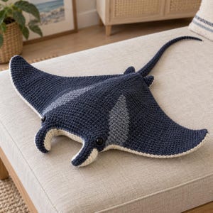 May include: A dark navy blue crochet manta ray plush toy featuring a long tail, two black plastic eyes, and light cream-colored accents on the underside and front fins. The textured yarn construction creates a soft, detailed marine animal figure.