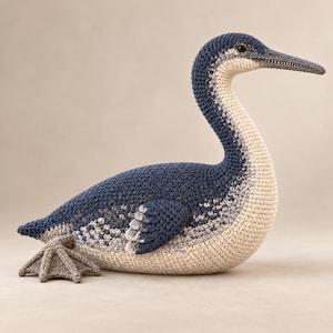 May include: A handcrafted crochet bird figurine featuring a long, slender neck and a pointed beak. The body displays a gradient of dark blue, grey, and cream-colored yarn. It has a single visible webbed foot in a light grey tone and a dark eye.