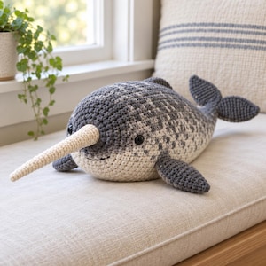 May include: A handmade crochet narwhal plush toy featuring a mottled grey and white body, a long cream-colored spiraled tusk, dark grey fins, and a dark grey tail. The narwhal has two small black plastic eyes and a stitched mouth on its face.