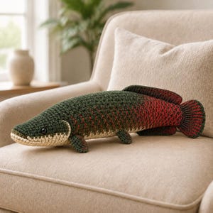 May include: A crochet fish plush toy featuring a dark green back that transitions into a deep red tail. The underside is a light cream colour, and the body has a textured, scale-like stitch pattern. It has a small black eye and fins.