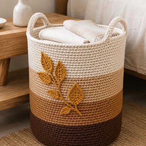 May include: A cylindrical crochet storage basket featuring a colour-blocked design in cream, tan, ochre, and dark brown. The basket has two integrated handles and is decorated with a prominent, textured, mustard-yellow crocheted leaf branch.