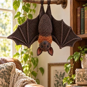 May include: A crocheted bat hanging upside down from a wooden branch. The bat features a dark brown body and wings, with a contrasting orange ruff of fur around its neck. It has pointed ears with pinkish inner details and small black eyes.