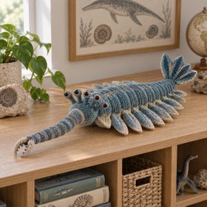 Könnte beinhalten: Ein gehäkeltes Plüschtier eines Anomalocaris, eines ausgestorbenen marinen Arthropoden. Das Wesen hat einen segmentierten Körper in Blau und Beige, einen langen vorderen Fortsatz mit klauenartigen Spitzen, mehrere seitliche Schwimmlappen und fünf Stielaugen.