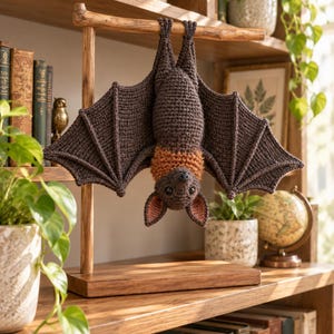 May include: A crocheted bat toy hanging upside down from a wooden T-shaped stand. The bat has dark brown wings and body, with a textured orange collar around its neck, black eyes, and pink-lined ears. The stand features a rectangular base.