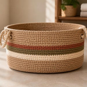 May include: A round, woven storage basket made of tan cord with two thick rope handles on the sides. The basket features horizontal stripes in muted terracotta, olive green, and cream colors encircling the middle section of the exterior.
