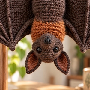 May include: A crocheted bat toy hanging upside down, featuring dark brown wings and body, a lighter orange-brown chest, and pink-toned inner ears. The bat has large, glossy black eyes and a small black nose, crafted with visible yarn stitches.