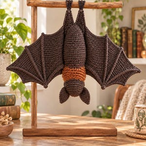 May include: A dark brown crocheted bat with a textured orange band around its midsection, hanging upside down from a wooden T-shaped display stand. The bat features detailed wing structures with visible stitching and two pointed ears.