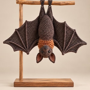 May include: A crocheted bat toy with dark brown wings and body, featuring a lighter orange-brown neck ruff and inner ears. The bat is hanging upside down from a wooden T-shaped display stand with a rectangular base, showcasing detailed yarn stitching.