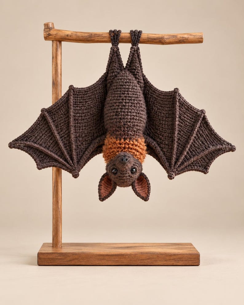 May include: A crocheted bat toy with dark brown wings and body, featuring a lighter orange-brown neck ruff and inner ears. The bat is hanging upside down from a wooden T-shaped display stand with a rectangular base, showcasing detailed yarn stitching.