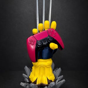 May include: A yellow and blue hand sculpture featuring three long silver claws extending upward, holding a bright pink and black video game controller. The base of the sculpture is surrounded by jagged gray rock-like formations.