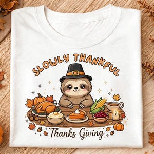 Könnte beinhalten: Ein weißes T-Shirt mit einer Grafik eines Faultiers mit Pilgerhut an einem Tisch voller Thanksgiving-Essen, darunter ein Truthahn, Kürbiskuchen und Mais. Der Text lautet SLOWLY THANKFUL und Thanks Giving in brauner Schrift.