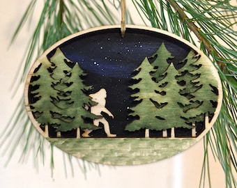 May include: A wooden Christmas ornament featuring a silhouette of Bigfoot walking through a forest at night. The ornament is painted with a dark blue background and green trees.