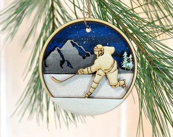 May include: A wooden Christmas ornament featuring a hockey player skating on a snowy field with mountains in the background. The sky is blue with white stars.