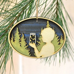 May include: Wooden oval ornament with a silhouette of a child holding a jar of fireflies against a dark blue and green forest background.