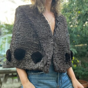 Pu&ograve; includere: Bolero in finta pelliccia marrone scuro e strutturata con tre applicazioni floreali nere sulle maniche. La giacca ha uno scollo a V e viene indossata sopra jeans blu. Stile vintage.