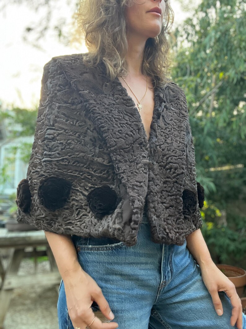 Pu&ograve; includere: Bolero in finta pelliccia marrone scuro e strutturata con tre applicazioni floreali nere sulle maniche. La giacca ha uno scollo a V e viene indossata sopra jeans blu. Stile vintage.