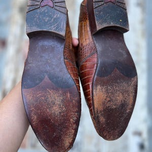May include: A pair of brown leather shoes with a textured, crocodile-like pattern. The soles are dark brown with wear and tear, showing the shoes have been used. The heels have a sunburst design.