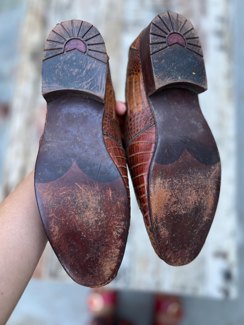 May include: A pair of brown leather shoes with a textured, crocodile-like pattern. The soles are dark brown with wear and tear, showing the shoes have been used. The heels have a sunburst design.